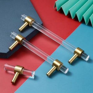 Pode incluir: Três botões de armário de acrílico transparente com detalhes em metal dourado. Os botões são de tamanhos diferentes e estão dispostos sobre uma superfície vermelha e azul.