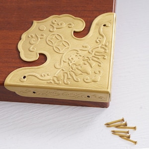 May include: Gold metal corner bracket with an ornate design. The bracket is attached to a wooden surface with screws. A set of small gold nails are included.