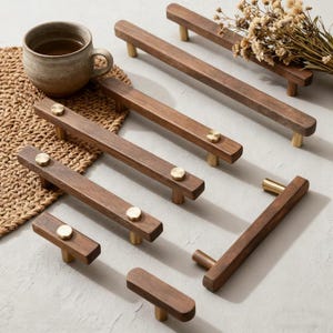 Pode incluir: Uma coleção de puxadores de armário de madeira com detalhes em latão. Os puxadores têm vários tamanhos e formatos, com um acabamento castanho escuro. Uma caneca de cerâmica com chá e flores secas também está na imagem. Para móveis e decoração.