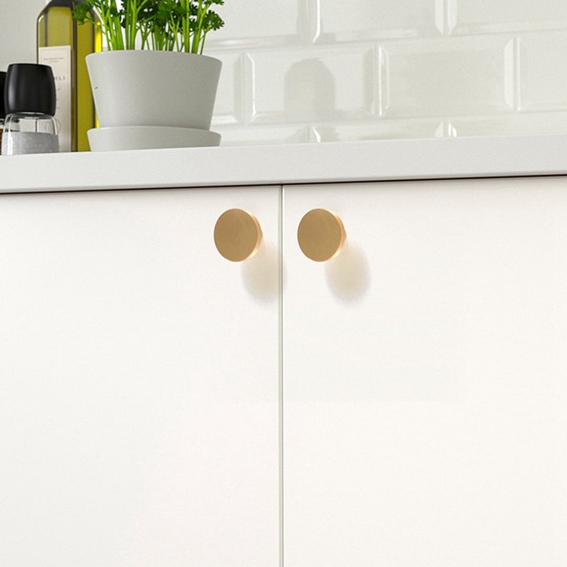 May include: Two round, gold cabinet knobs are mounted on a white cabinet door.