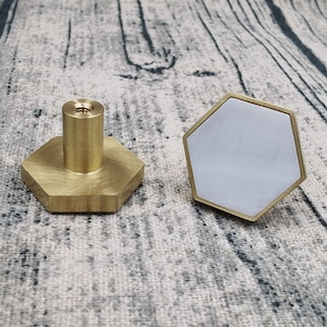 May include: Two hexagonal cabinet knobs with a brass finish. One knob has a white center, while the other has a brass center.