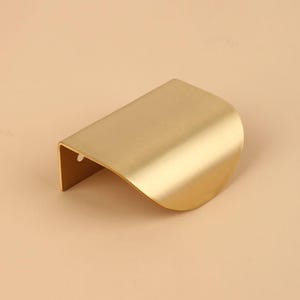 May include: A gold-coloured, curved cabinet pull with a brushed finish. The handle has a rounded, wave-like shape and a rectangular base. This hardware is designed for easy installation and adds a modern touch to furniture.