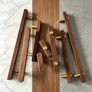 May include: A collection of cabinet pulls featuring dark brown wood bars and cylindrical brass metal accents. The hardware includes various lengths of rectangular wooden handles with gold-toned circular mounting points and cylindrical posts.