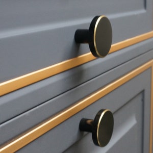 May include: Two black cabinet knobs with a gold trim on a grey cabinet with gold trim.