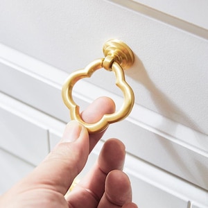 May include: Gold-toned metal drawer pull with a four-leaf clover design. The pull is attached to a white cabinet door.