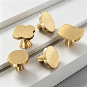 May include: A collection of five cabinet knobs in a brushed gold finish. The set includes two circular knobs, one clover-shaped knob, one square knob with rounded edges, and one cylindrical knob, all featuring a smooth, metallic surface texture.