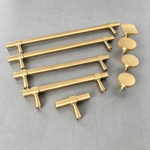 May include: A collection of brushed gold cabinet hardware including four horizontal bar pulls of varying lengths, one small T-shaped knob, and four circular knobs. Each piece features a smooth, matte metallic finish and cylindrical design.
