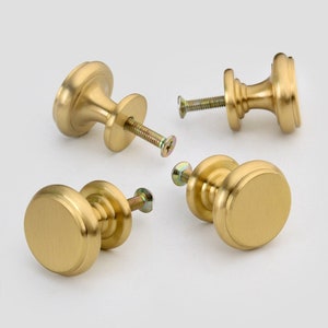 May include: Four gold-toned cabinet knobs with a round, slightly raised, flat top and a rounded base. Each knob has a screw for installation.