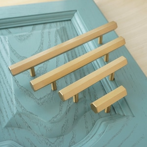 May include: Four gold-toned cabinet pulls with a hexagonal shape. The pulls are arranged in a row, with the largest pull at the top and the smallest pull at the bottom.