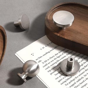 Peut inclure: Boutons de meubles en nickel brossé de différentes formes : un disque plat, une sphère et une base cylindrique avec un dessus plat. Les boutons sont présentés sur une surface grise avec un plateau en bois et une page de texte d'un livre.