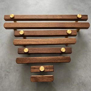 Può includere: Una collezione di sette maniglie per cassetti in legno disposte a triangolo. Ogni maniglia presenta una barra rettangolare in legno marrone scuro con elementi di fissaggio circolari in ottone spazzolato alle estremità o al centro.