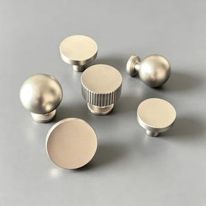 May include: A collection of six silver-toned metal cabinet knobs with a brushed finish. The set includes four flat-topped circular knobs, one spherical knob, and one cylindrical knob featuring a vertically ribbed texture around its side edge.
