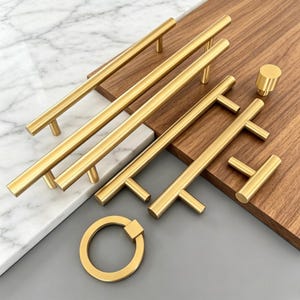 May include: A collection of brushed gold cabinet hardware including four long cylindrical bar pulls, one small T-shaped handle, one cylindrical knob, and one circular ring pull. The hardware features a smooth, matte metallic finish.