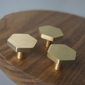 Könnte beinhalten: Drei goldene sechseckige Schrankknöpfe mit gebürsteter Oberfläche. Die Knöpfe befinden sich auf einer Holzoberfläche.