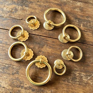 May include: A collection of seven gold-toned metal ring pulls arranged in a circular pattern. Each pull features a smooth circular ring attached to a decorative, scalloped, flower-shaped backplate with a central mounting point for furniture hardware.