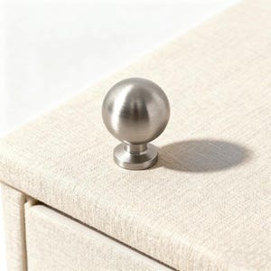 May include: A brushed nickel sphere knob with a circular base, mounted on a light beige textured surface. The knob casts a shadow, highlighting its spherical shape. This hardware is suitable for drawers, cabinets, or furniture.
