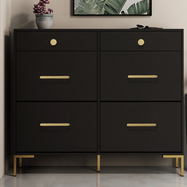 May include: A black dresser with six drawers and gold-toned hardware. The dresser has four legs and sits on a light-colored floor.