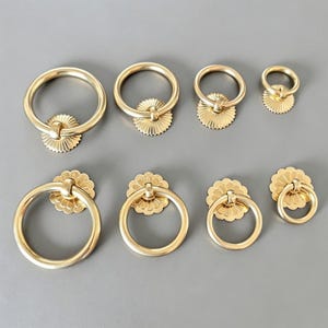 Könnte beinhalten: Eine Sammlung von acht goldfarbenen Metallringgriffen, die in zwei Reihen angeordnet sind. Jeder Griff besteht aus einem kreisförmigen Ring, der an einer dekorativen, muschel- oder sonnenförmigen Rückplatte befestigt ist. Die Größen variieren von groß bis klein.