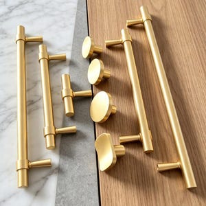 May include: A collection of brushed gold cabinet hardware including long cylindrical bar pulls, T-shaped handles, and various round and contoured knobs. The hardware features a smooth, matte metallic finish and simple, clean geometric shapes.