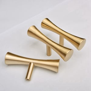 May include: Four gold-toned cabinet pulls arranged on a white surface. Each pull has a cylindrical stem and a curved, hourglass-shaped handle. The handles are smooth and reflect light, suggesting a polished finish. These pulls are designed for furniture.