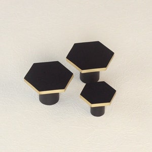 Pode incluir: Três botões hexagonais pretos para armário com acabamento dourado. Os botões são de tamanhos diferentes e estão dispostos em forma escalonada.