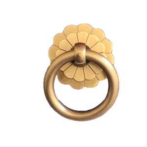Könnte beinhalten: Ein goldfarbener Ring-Schrank-Knopf mit einer dekorativen, blütenförmigen Rückplatte.