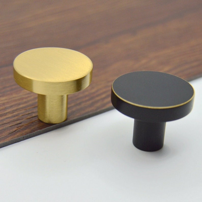 May include: Two round cabinet knobs, one gold and one black, with a simple, modern design. The knobs are made of metal and have a smooth, polished finish.
