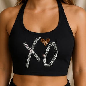 May include: Black halter crop top with a ribbed texture. The top features the letters "XO" embellished with rhinestones, and a small heart made of gold-colored rhinestones. A stylish and trendy top.
