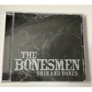 May include: A CD case with a black and white image of a patterned fabric. The text "THE BONESMEN SKIN AND BONES" is printed in white on the front of the case.