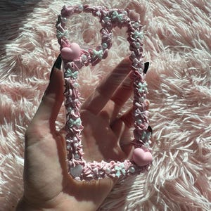 Puede incluir: Una funda de teléfono rosa con un diseño decorativo. La funda presenta un borde texturizado y elevado adornado con pequeños lazos blancos, perlas y corazones rosas. La funda se sostiene contra un fondo rosa esponjoso.