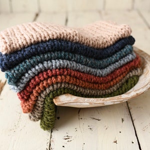 Può includere: Un mucchio di quadrati di tessuto a maglia in vari colori, tra cui beige, blu navy, verde acqua, grigio, ruggine, marrone e verde, disposti in una forma curva in una ciotola di legno bianca.