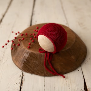 May include: Red knitted baby hat with a tie, on a wooden surface with red berries.