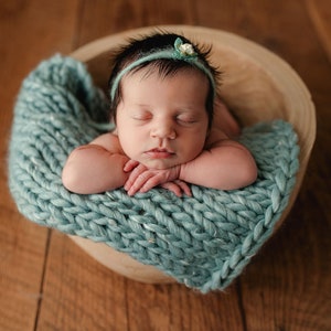 May include: A newborn baby sleeping peacefully in a wooden bowl lined with a teal knitted blanket. The baby is wearing a headband with a small white flower.
