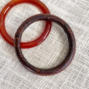 Op de afbeelding: Twee cirkelvormige armbanden. Eén is een doorschijnende rood-oranje kleur. De andere is donkerbruin met een textuur, reptielachtig patroon. De armbanden liggen op een lichtbeige, getextureerd oppervlak.