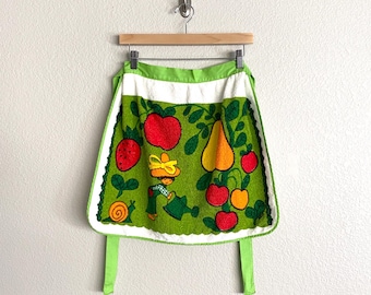Könnte beinhalten: Eine grüne Vintage-Schürze mit weißem Rand und lindgrünen Bindebändern. Die Schürze zeigt ein skurriles Design mit Früchten, einer Schnecke und einem Gärtner, der Pflanzen gießt. Die Schürze hängt an einem Holzbügel.