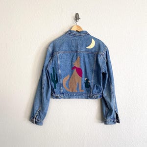 May include: A light-wash denim jacket with a desert-themed design. The back features a howling wolf patch with a red bandana, a gold crescent moon, and green cactus appliqués. The jacket is hanging on a wooden hanger.