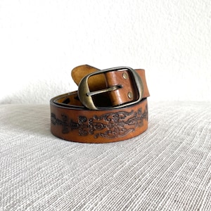 Puede incluir: Un cinturón de cuero marrón con un diseño en relieve ornamentado. El cinturón tiene una hebilla de latón y está enrollado. El patrón en relieve es visible en la superficie del cinturón, agregando un elemento decorativo.