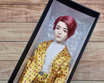May include: A doll with pink hair wearing a yellow and white patterned suit jacket and pants. The doll is lying in a black box with white packing peanuts.