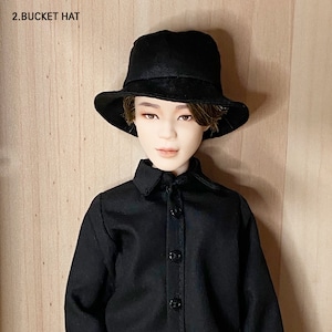 Può includere: Un cappello a secchio nero con una tesa leggermente inclinata verso il basso. Il cappello è indossato da una bambola vestita con una camicia nera abbottonata. Il testo "2. BUCKET HAT" è in alto a sinistra.