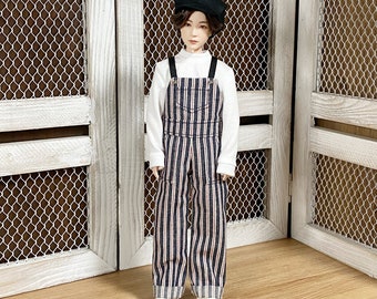 May include: A doll wearing a white turtleneck, striped overalls, a black cap, and white sneakers. The overalls have vertical stripes in navy, red, and white. The doll is standing in front of a white wooden door.