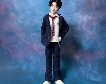 May include: A doll dressed in a navy blue suit with a white shirt and a red and white striped tie. The doll is wearing white sneakers and has black hair. A silver chain hangs from the doll's pants pocket. The background is a blue and purple gradient.