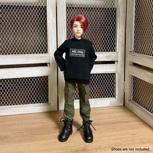 Può includere: Una bambola indossa un maglione nero con la scritta "MIC DROP How you think bout that" e pantaloni cargo verde oliva. La bambola ha i capelli rossi e stivali neri. Lo sfondo include una porta di legno bianca con una rete metallica.