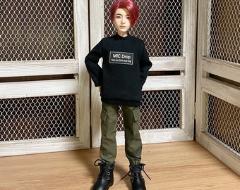 May include: A doll wearing a black sweater with the text "MIC DROP How you think bout that" and olive green cargo pants. The doll has red hair and black boots. The background includes a white wooden door with a wire mesh.