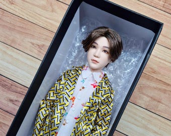 May include: A doll with brown hair wearing a yellow and black patterned jacket and a white shirt with floral print. The doll is lying in a black box with white packing peanuts.