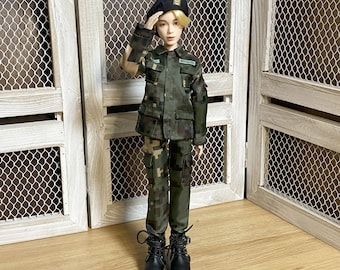 May include: A doll wearing a military uniform, including a green and brown camouflage jacket and pants. The doll is also wearing a black beret and black boots. The doll is saluting. The text in the image says: Shoes are not included. The pattern of the military uniform is slightly different from the photo.