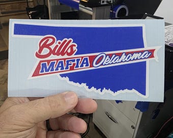 Puede incluir: Una pegatina blanca con el contorno azul de Oklahoma y las palabras "Bills Mafia Oklahoma" en rojo y blanco. La pegatina está sujeta en la mano de una persona.