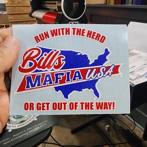 May include: A rectangular sticker with the text "RUN WITH THE HERD" in red arched letters. Below, "Bills" is in red script, and "MAFIA USA" is in white letters on a red banner over a blue map of the USA. The bottom text reads "OR GET OUT OF THE WAY!"