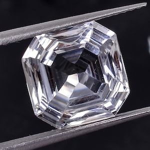 May include: A clear, faceted, square-shaped gemstone held by tweezers against a black background. The gemstone is a cut and polished crystal.