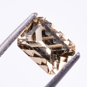 May include: A light brown gemstone with a rectangular shape, held by tweezers against a white background.