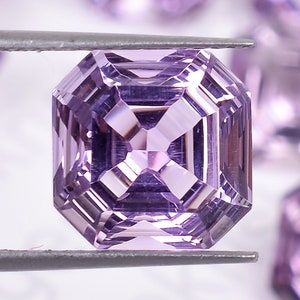 May include: A single, large, light purple, emerald-cut gemstone is held by tweezers. The gemstone is surrounded by other smaller, similar gemstones.
